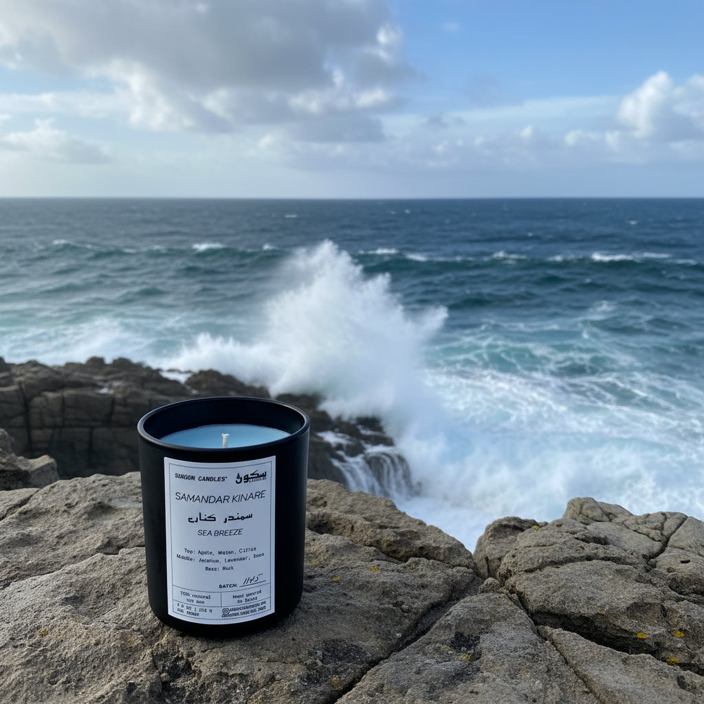 Candle in a black container with a white label on a rocky surface with waves crashing in the ocean in the background